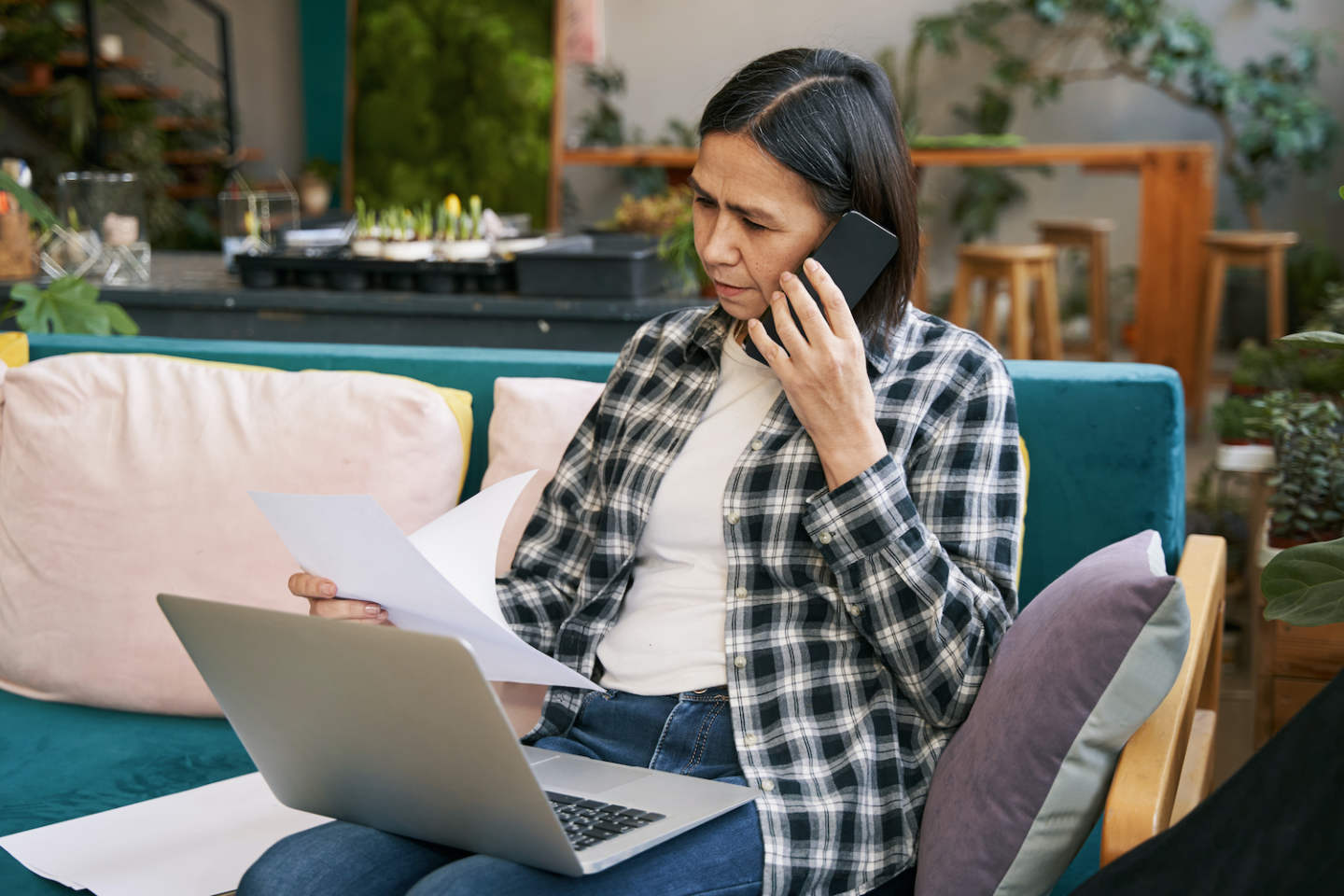 Female in plaid shirt sitting on the coach, working at laptop, holding papers and talking on the phone