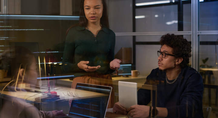 Young female programmer standing at table having discussion with her colleagues in modern office in evening