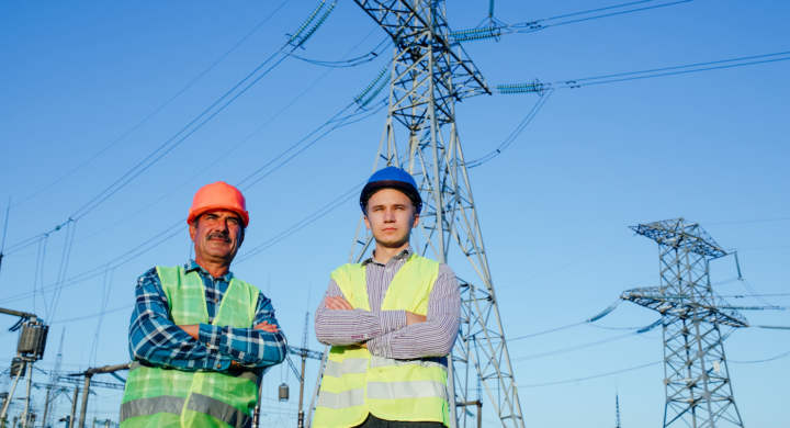 Low angle of confident serious professional male electricians technicians in hardhats and waistcoats looking away while standing together against blue sky and modern electric towers