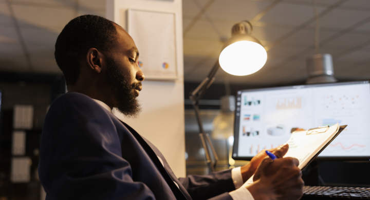 Careful entrepreneur analyzing marketing graphs, working late at night at company investment plan in startup office. African american worker planning strategy to help increase profit