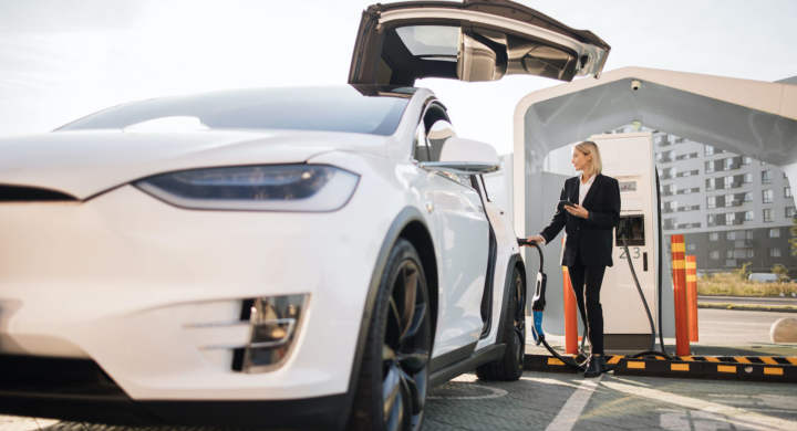 Beautiful caucasian woman in formal wear using modern smartphone while charging white lectro car at station. Concept of sustainable and economic transportation.