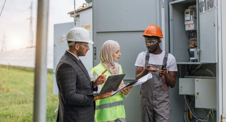 Indian man and muslim woman with clipboard and laptop talking with african american technician at solar station. Multiracial people examining process of green energy production.