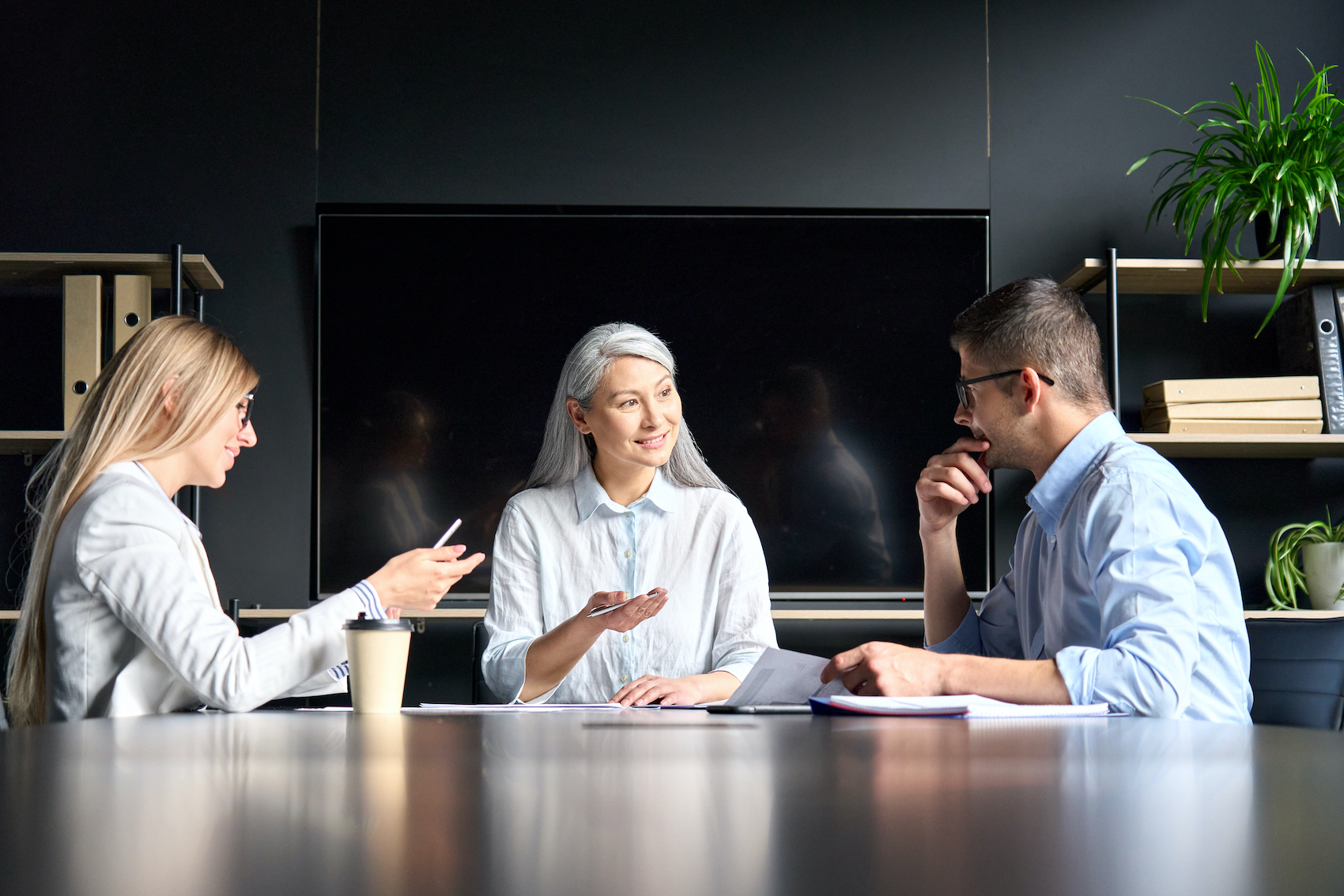 Senior mid aged Asian executive mentor teacher talking to younger male and female Caucasian colleagues trainees interns students on business seminar training in meeting room with big screen monitor.