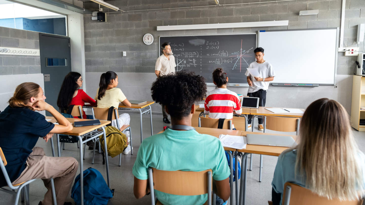 Teen asian boy high school student giving a presentation in class to his multiracial classmates and male teacher. Horizontal banner image.