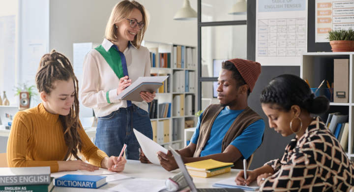 Young smiling teacher conducting foreign language lesson with students while they sitting at table with books and computer