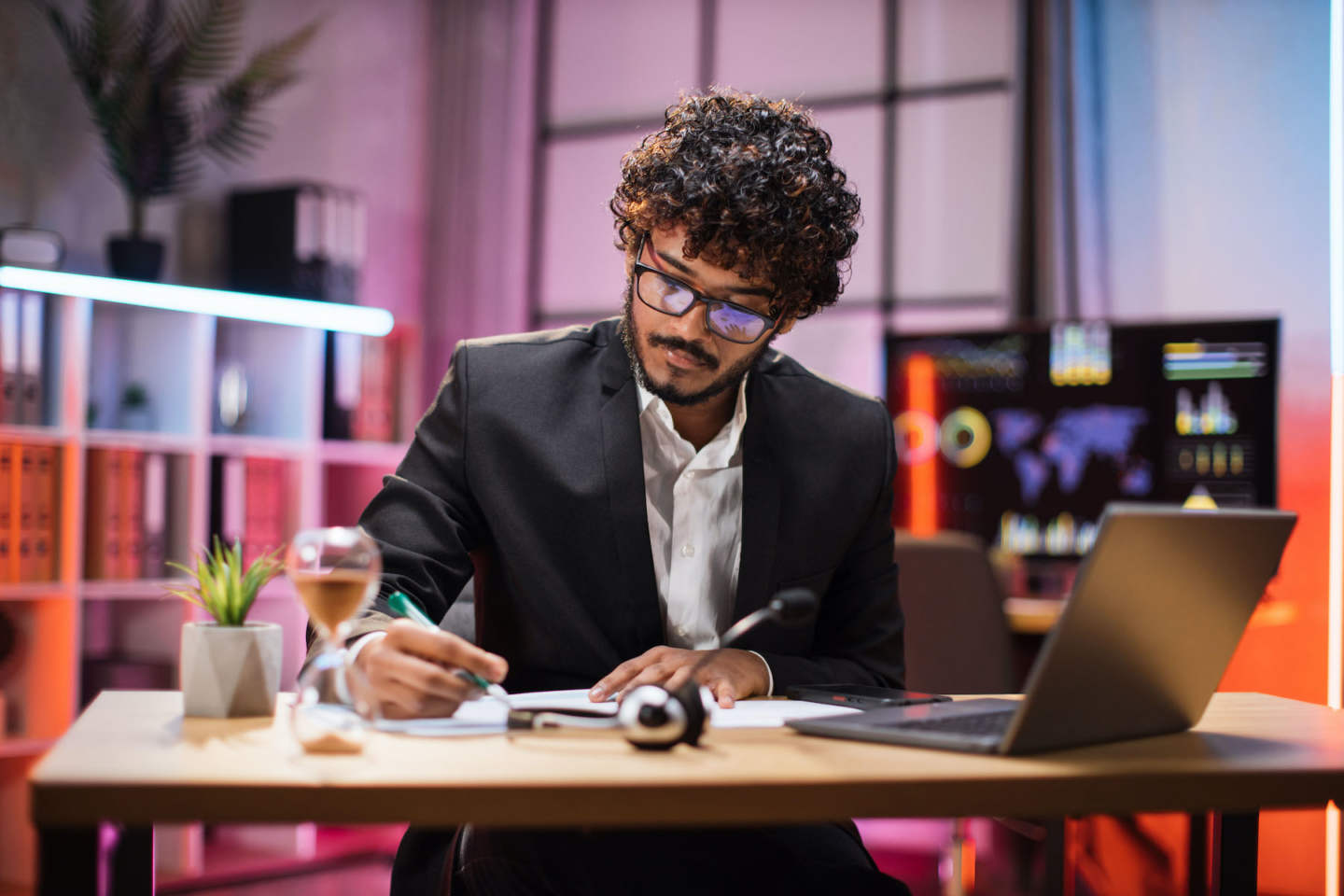 Attractive confident bearded businessman, office manager, wearing suit using laptop while making, writing financial report, using pen, on paper working at night.
