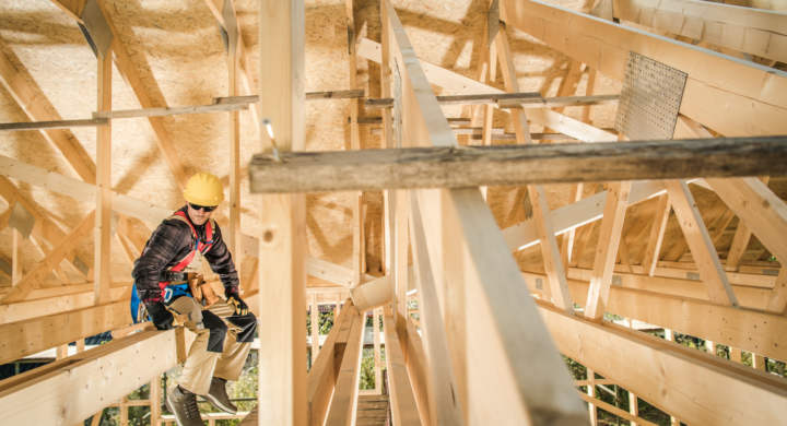 Caucasian Contractor in His Work Wear and Yellow Construction Safety Helmet Taking a Break During Work, Resting on a Roof Beam of a Newly Built Wooden House Skeleton. Construction Site Theme.
