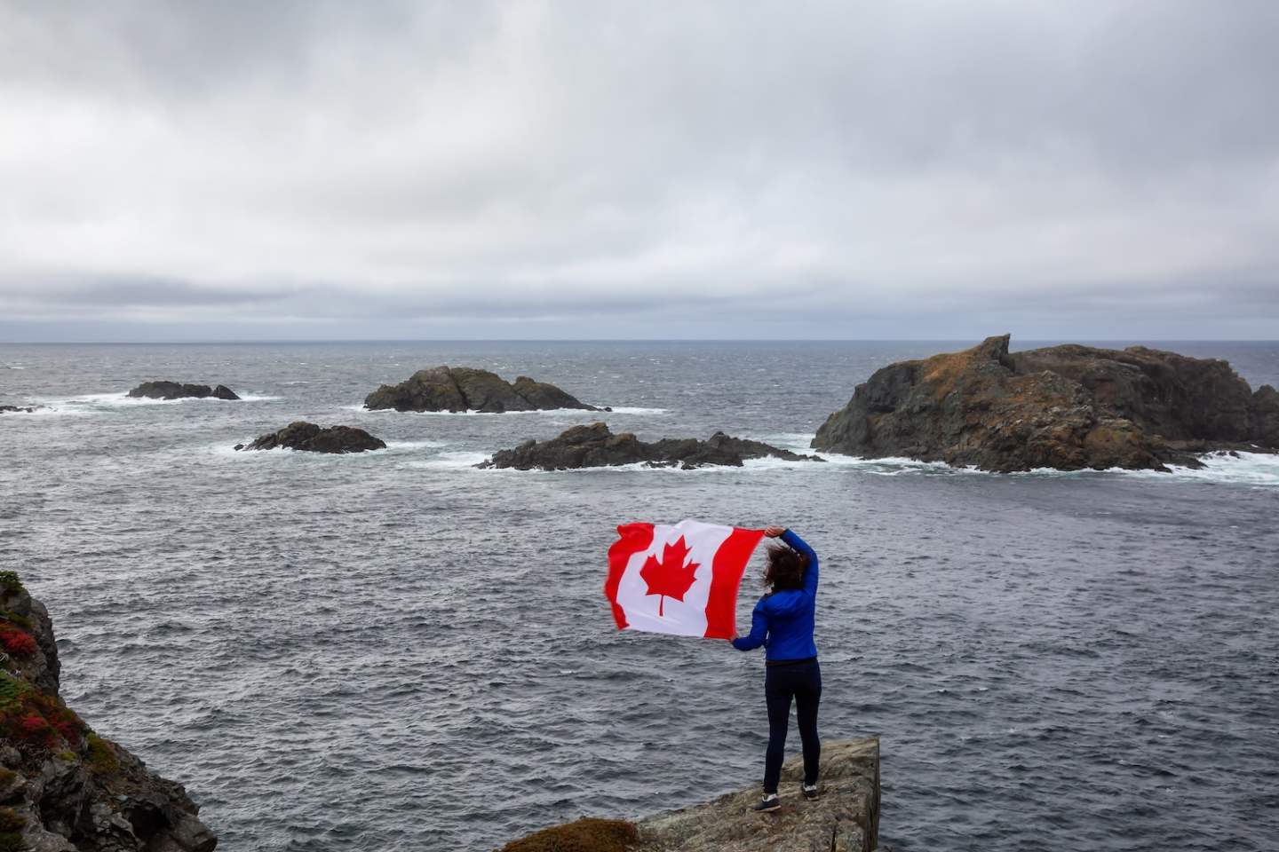 Adventurous woman holding a Canadian Flag on a Rocky Atlantic Ocean Coast during a cloudy day. Taken in Sleepy Cove, Crow Head, Twillingate, Newfoundland, Canada.