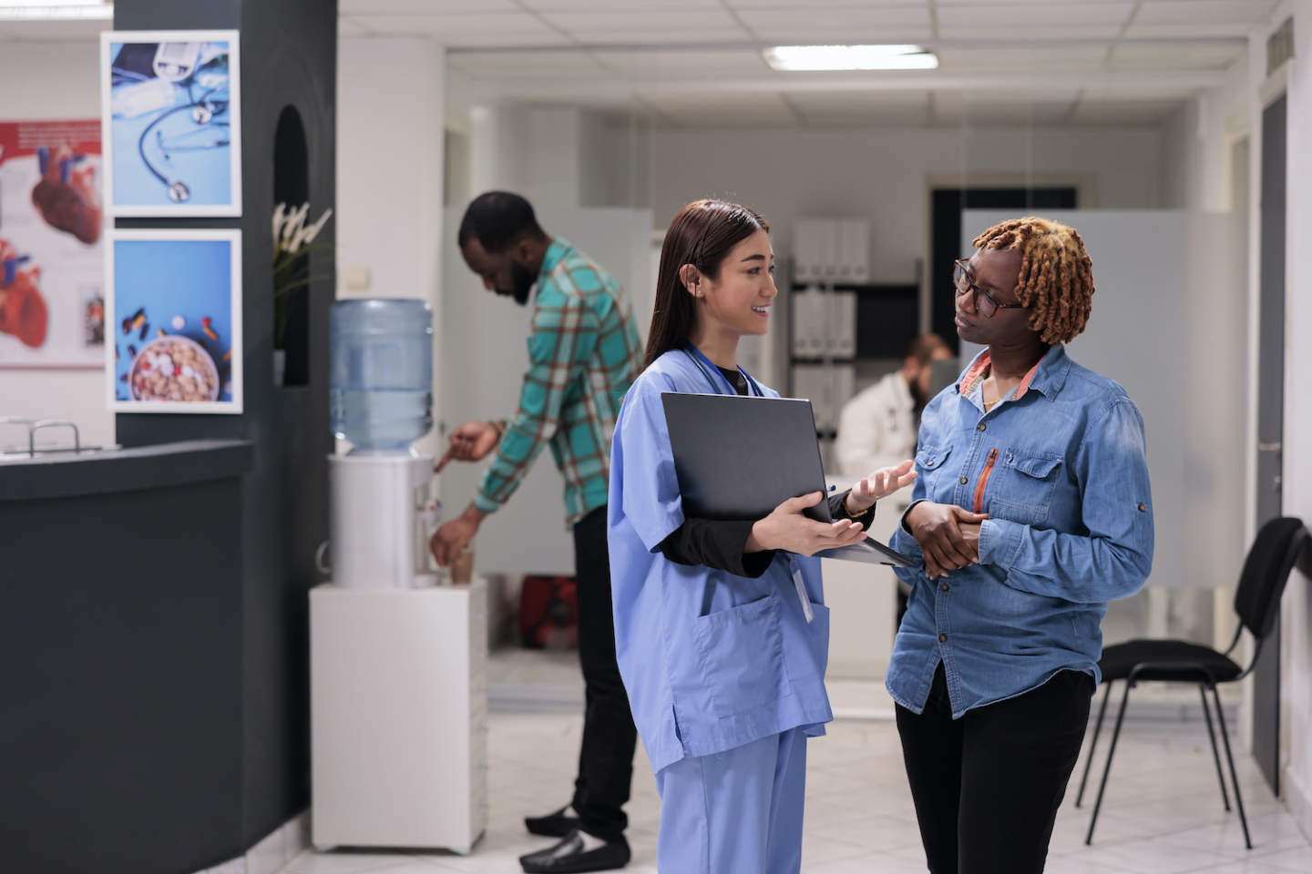 Asian nurse showing disease diagnosis to person in hospital reception lobby, explaining medical results on laptop. Diverse women talking about medicine treatment and recovery in waiting area.