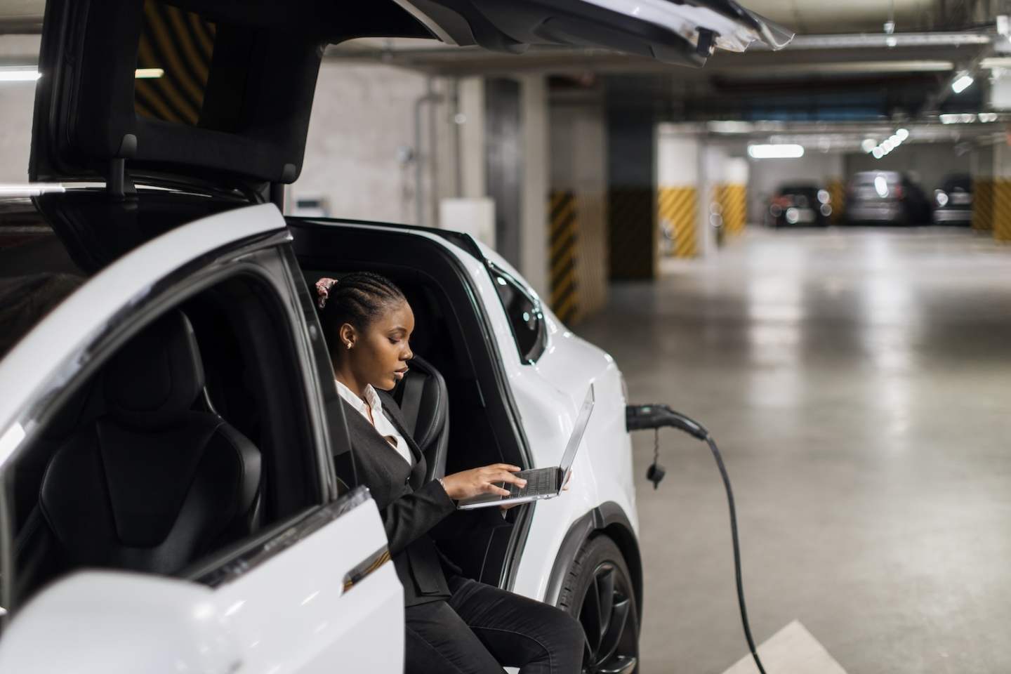 Side view of african businesswoman typing on portable computer while sitting in electric car in below-level parking lot. Confident office worker checking digital reports while auto being refilled.