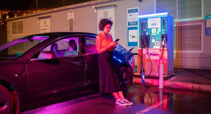 Girl with smartphone standing on wet asphalt by electric car during charging process