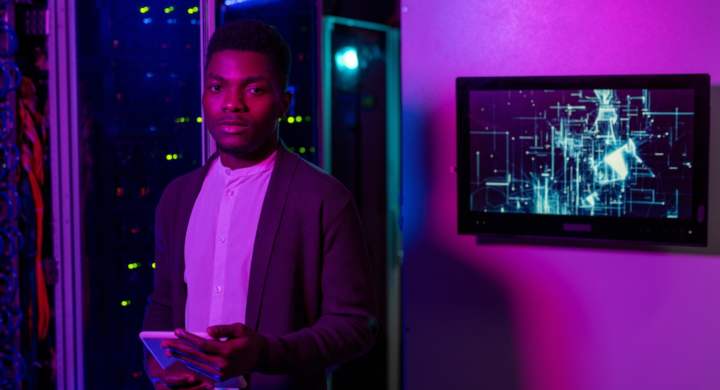 Portrait of confident young Black IT engineer in cardigan standing with tablet in dark neon-lighted room and working with supercomputer