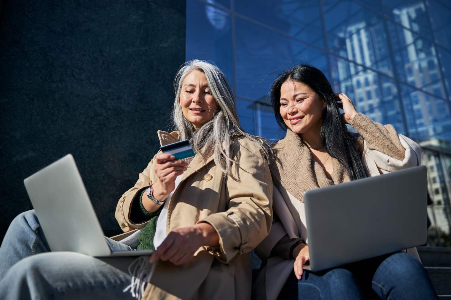 Charming lady with notebook on her laps holding credit card and smiling while spending time with friend outdoors