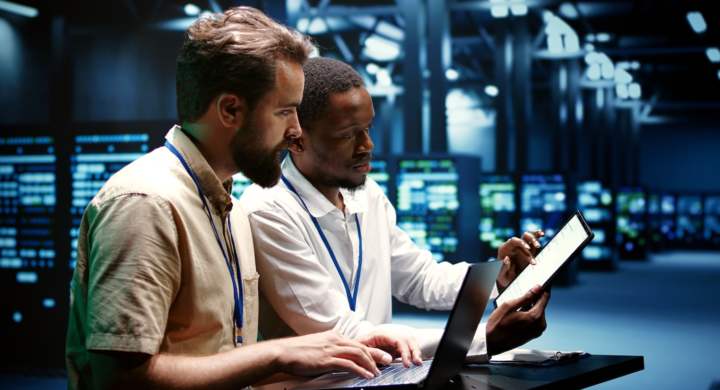 Coworkers running data center hardware diagnostic tests to determine and service software issues. Servicemen using laptop and tablet to fix critical systems, checking for blips