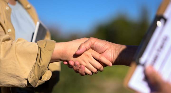 Cropped shot of farmers shaking hands at the field against blue sky. Agreement and contract in agriculture.
