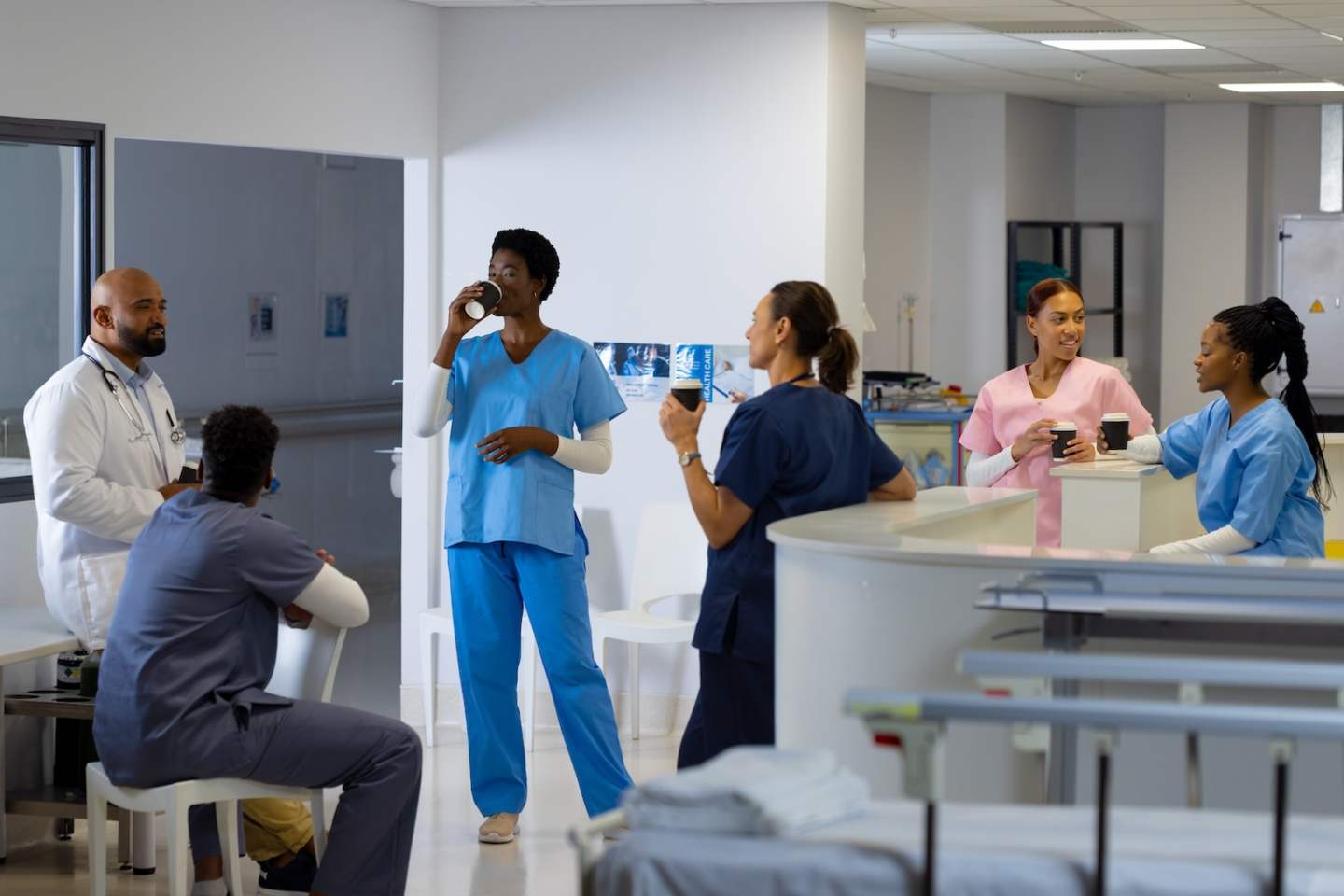 Happy diverse doctors wearing scrubs drinking coffee and talking at reception desk at hospital. Hospital, teamwork, medicine, healthcare and work, unaltered.