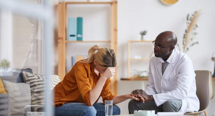 Serious young bald mental health doctor in white coat holding hand of patient while giving psychological support to her during therapy session
