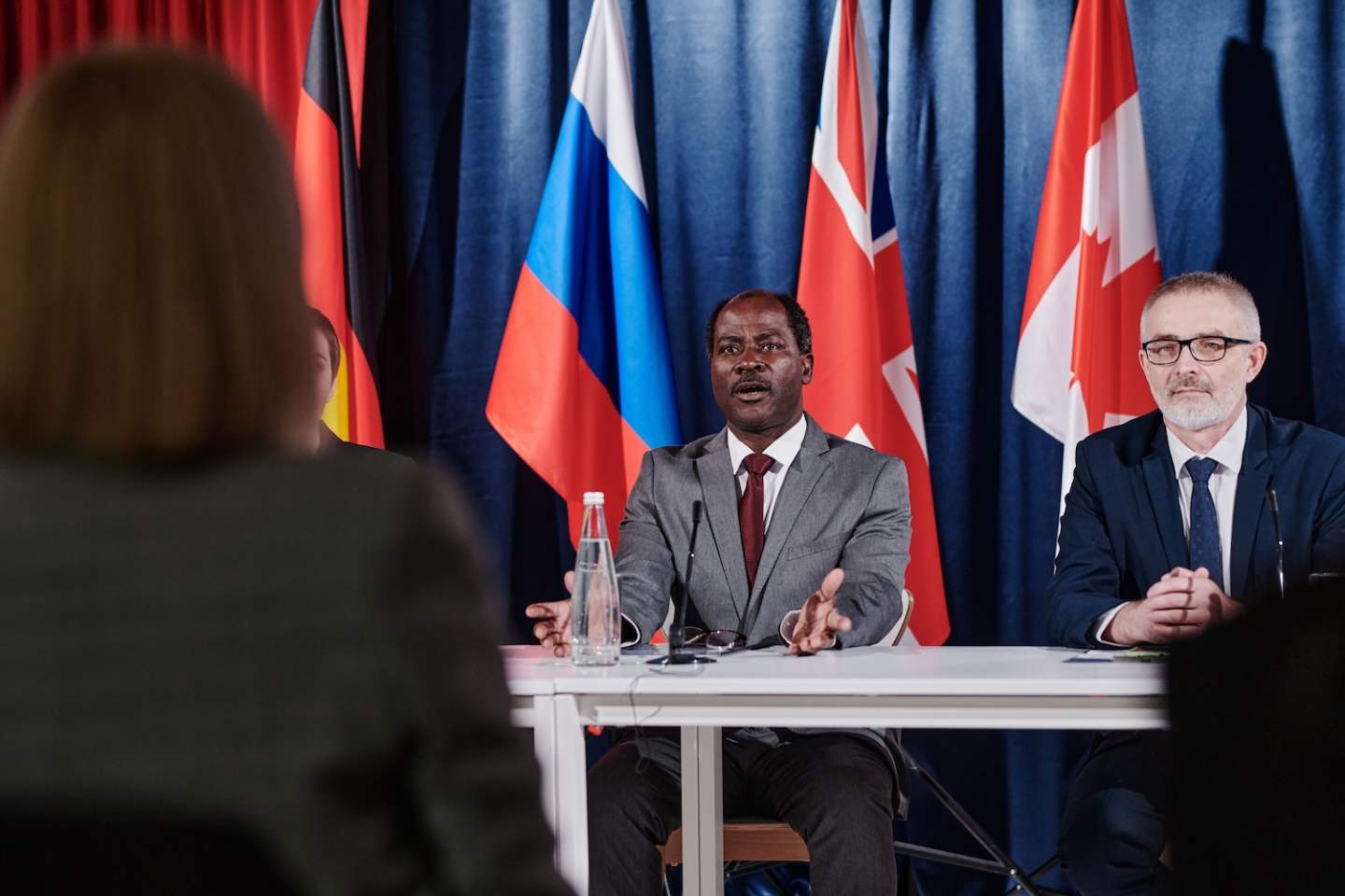 African male politician in suit sitting at table with his colleagues and answering questions asking by journalist at press conference