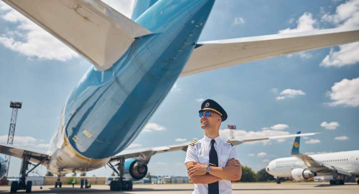 Handsome young man airline worker crossing arms and smiling while standing outdoors with airplanes and sky on background
