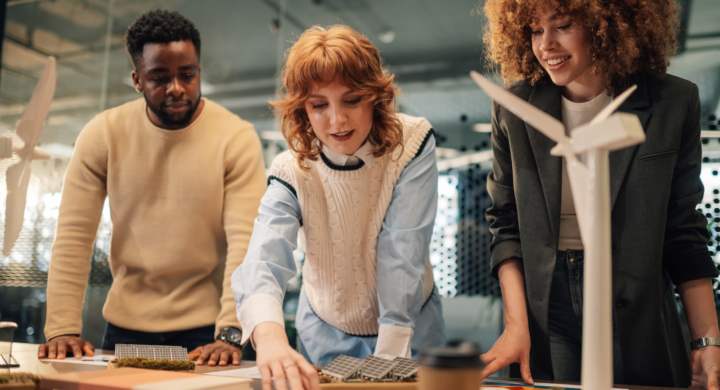 Multicultural businesspeople standing at modern workspace with windmill and solar panels model on table and analyzing charts for renewable energy solution. Staff working on clean energy technology