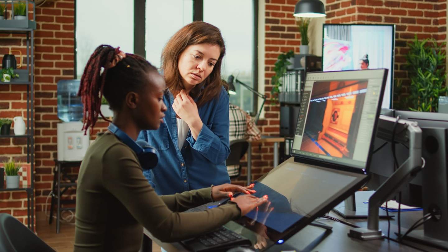 Diverse colleagues analyzing game developing electronics and new production interface in creative agency office. Artistic engineer talking to coworker about professional software creation.