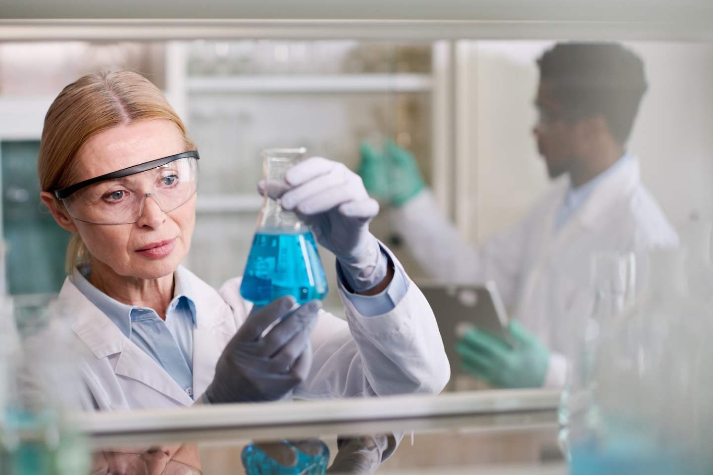 Medium close up shot of senior female medical laboratory scientist in safety glasses holding conical flask with liquid substance in gloved hands, male colleague working in blurred background