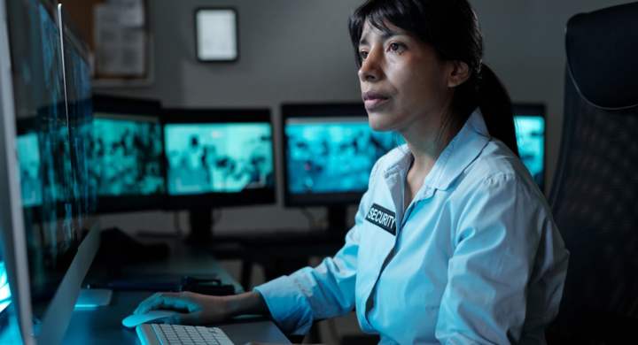 Young serious security guard in uniform sitting by desk in control room and looking at desktop computer screen while clicking mouse