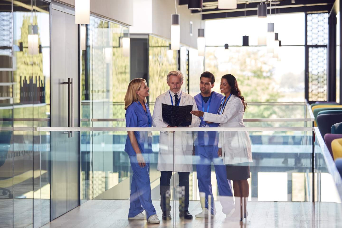 Multi-Cultural Medical Team Wearing White Coats And Scrubs Discuss Patient Scan Meeting In Hospital