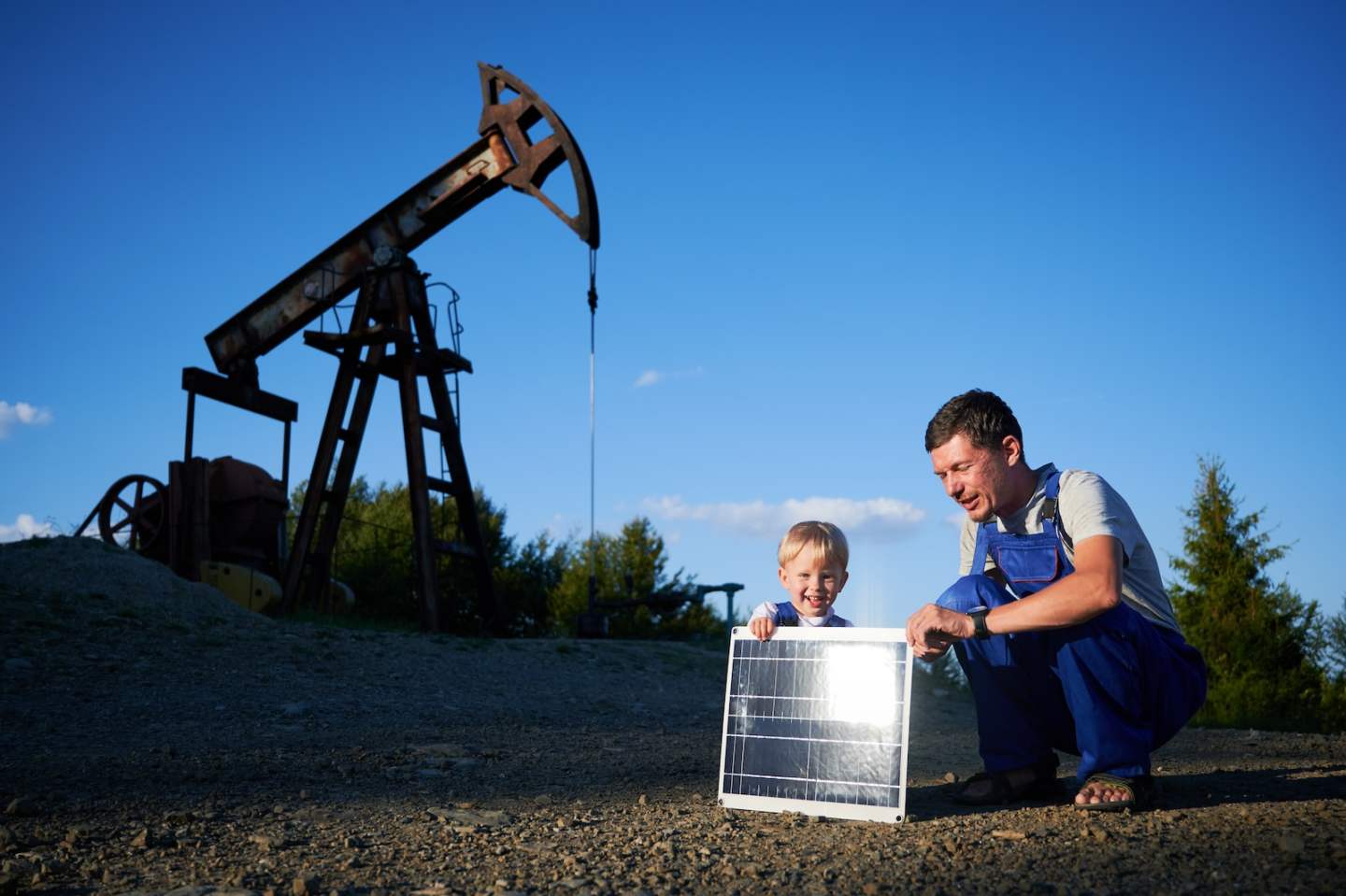 Adult man and male kid sat down on gravel site near oil well. They holding solar battery, sun rays reflected from solar panel. Two variant of energy production with different impact on environment.