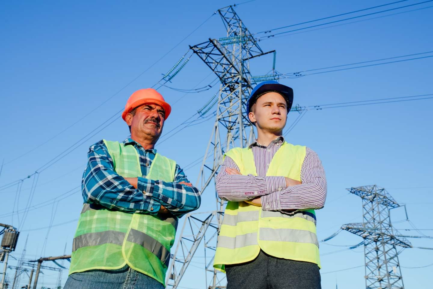 Low angle of confident serious professional male electricians technicians in hardhats and waistcoats looking away while standing together against blue sky and modern electric towers