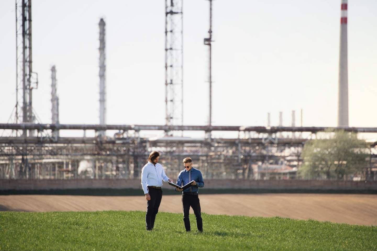 Two young engineers standing outdoors by oil refinery, discussing issues. Copy space.