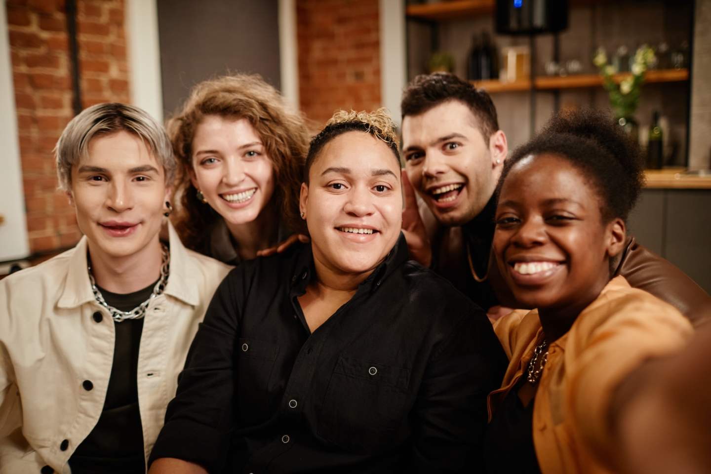Group of young cheerful friends posing for selfie in front of camera while relaxing on couch in living room at home party on weekend