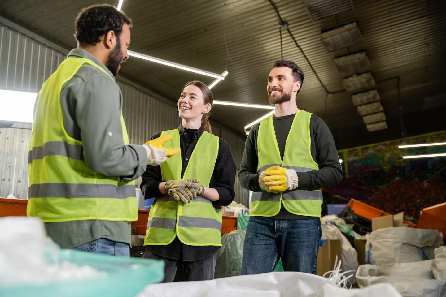 Bearded indian worker in protective glove and vest talking on cheerful colleagues while standing together near blurred sacks with trash in waste disposal station, recycling concept