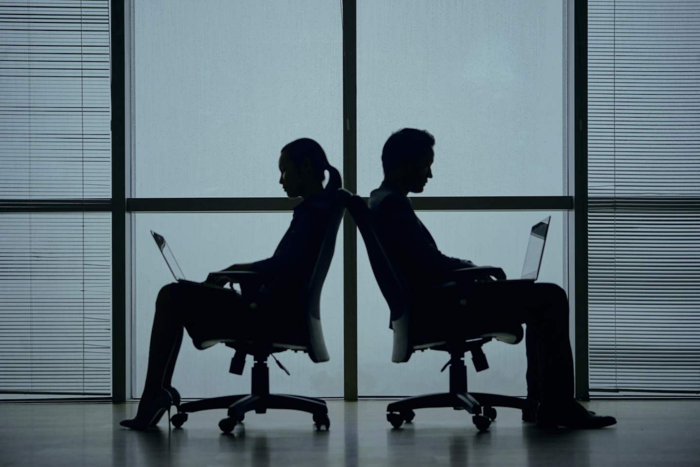 Silhouettes of two business people sitting back to back in office chairs against big panoramic window and working on laptop