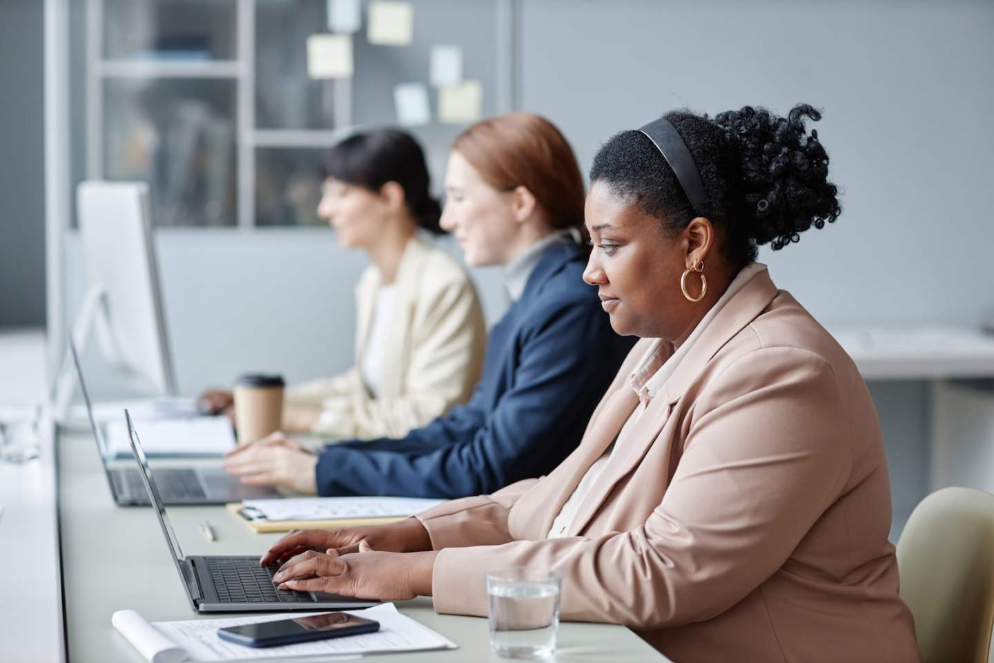 Side view of adult African American female company employee wearing pastel suit jacket typing on laptop while sitting at desk with coworkers in blurred background