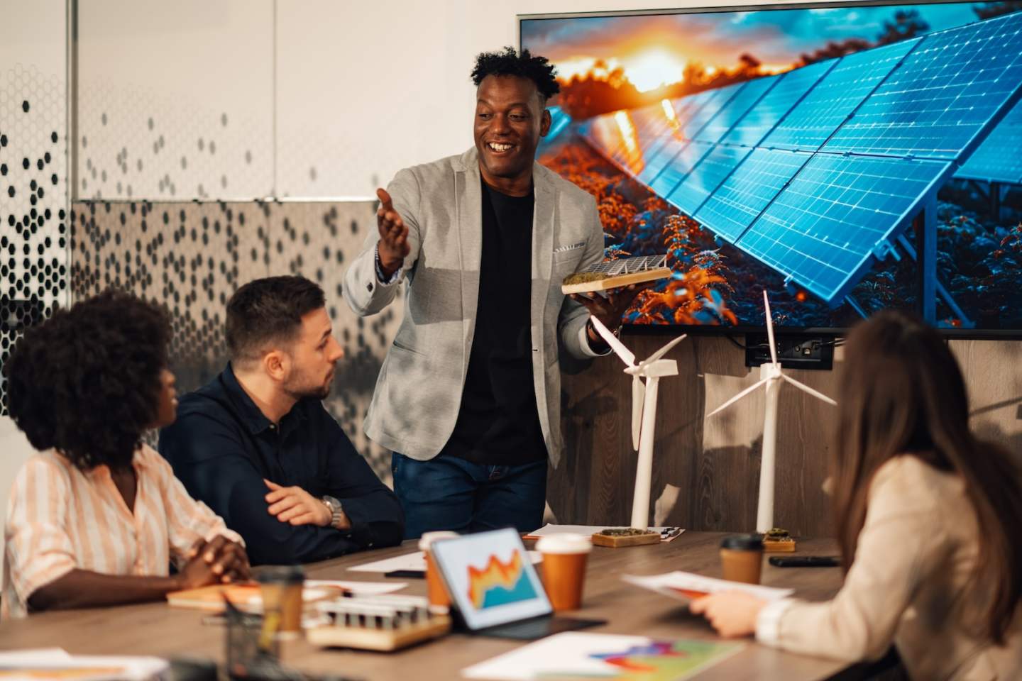 Smiling multicultural ecologist standing at conference room with solar panel miniature in hands and having meeting with eco staff during meeting. Interracial businesspeople working on climate change.