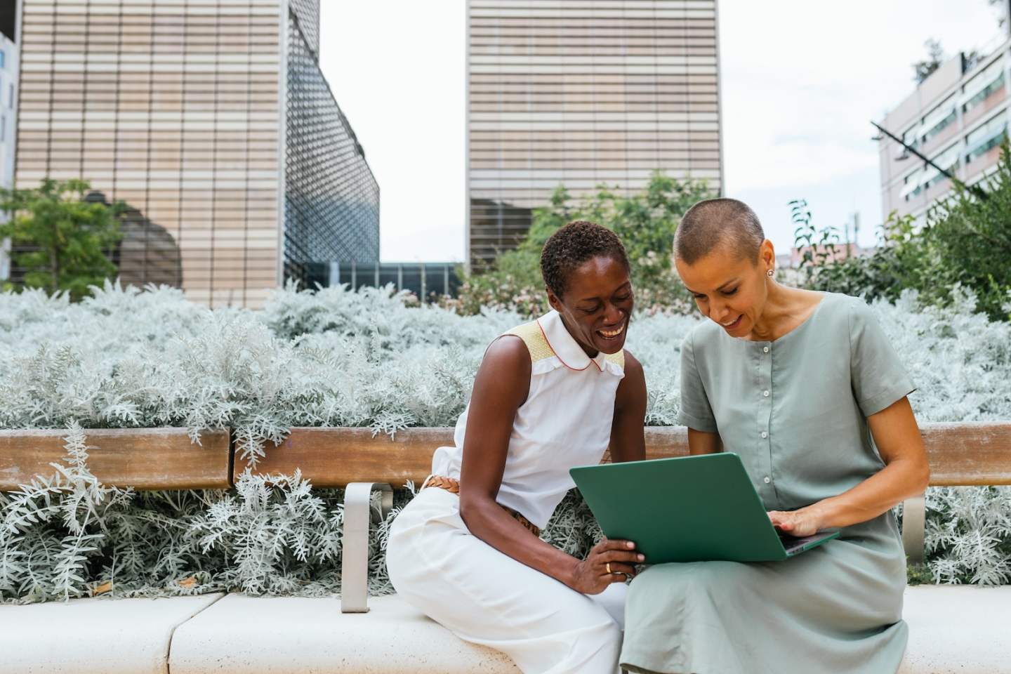 two businesswomen working with laptop and talking in park in front of business buildings