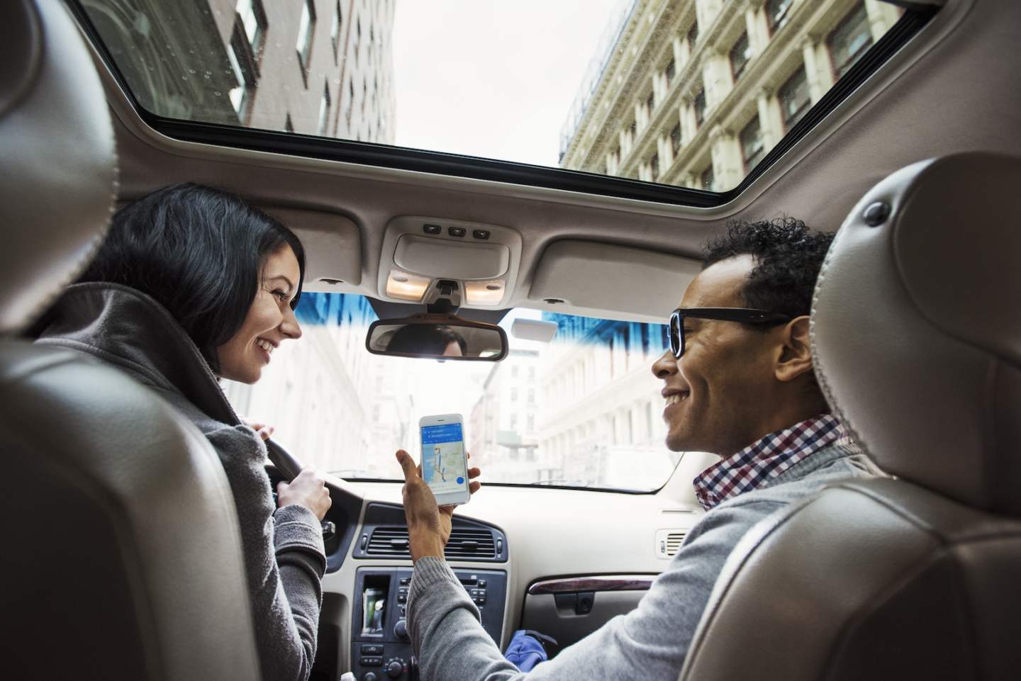 A young woman and young man in a car looking at a map on the display of a cellphone, seen from the back seat.