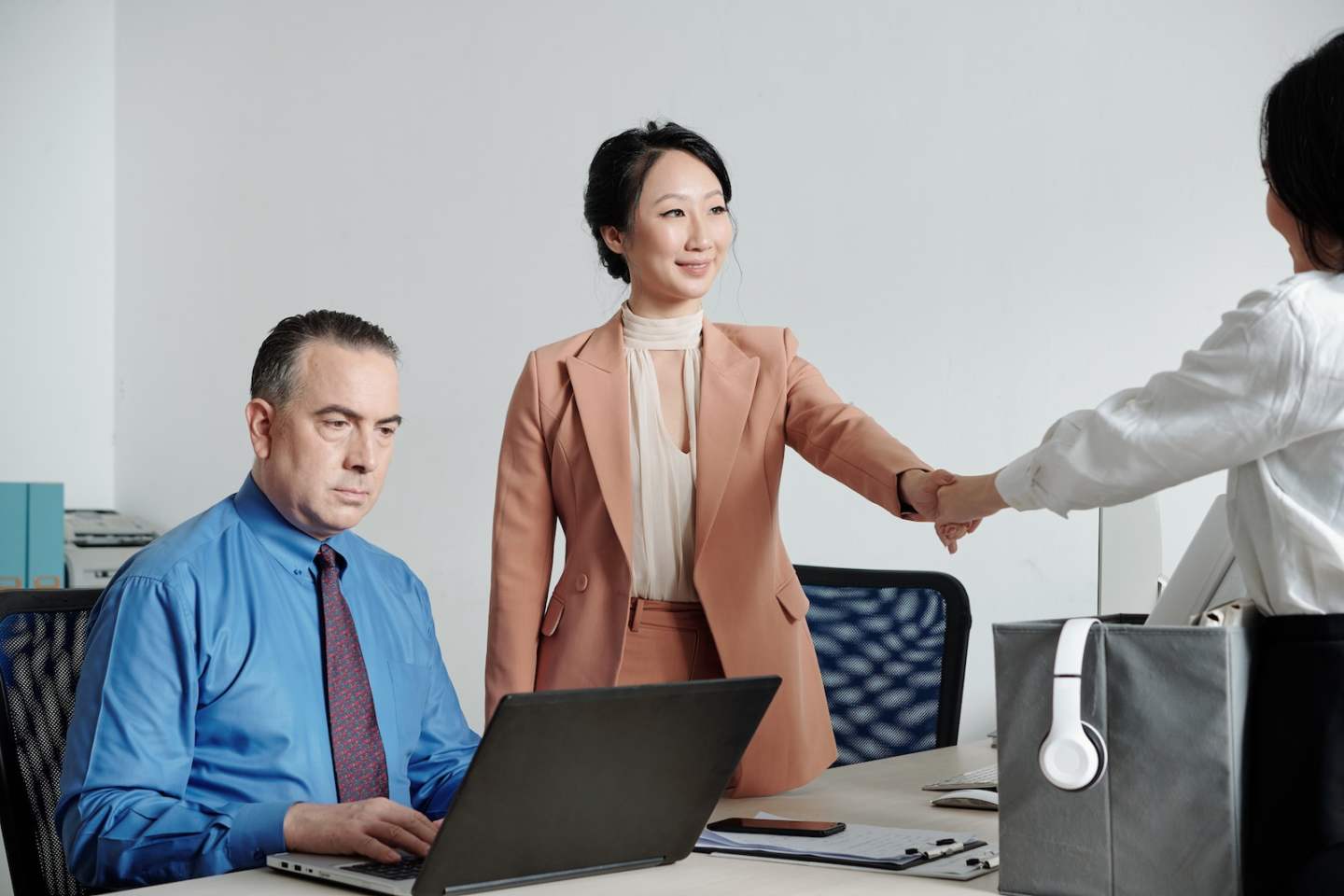Serious mature entrepreneur working on laptop, his colleague greeting and shaking hand of new office worker