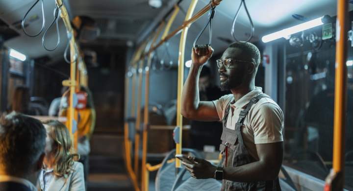 An African American worker in overalls standing on a night bus holding a handrail and using a smart phone in a bus full of people.