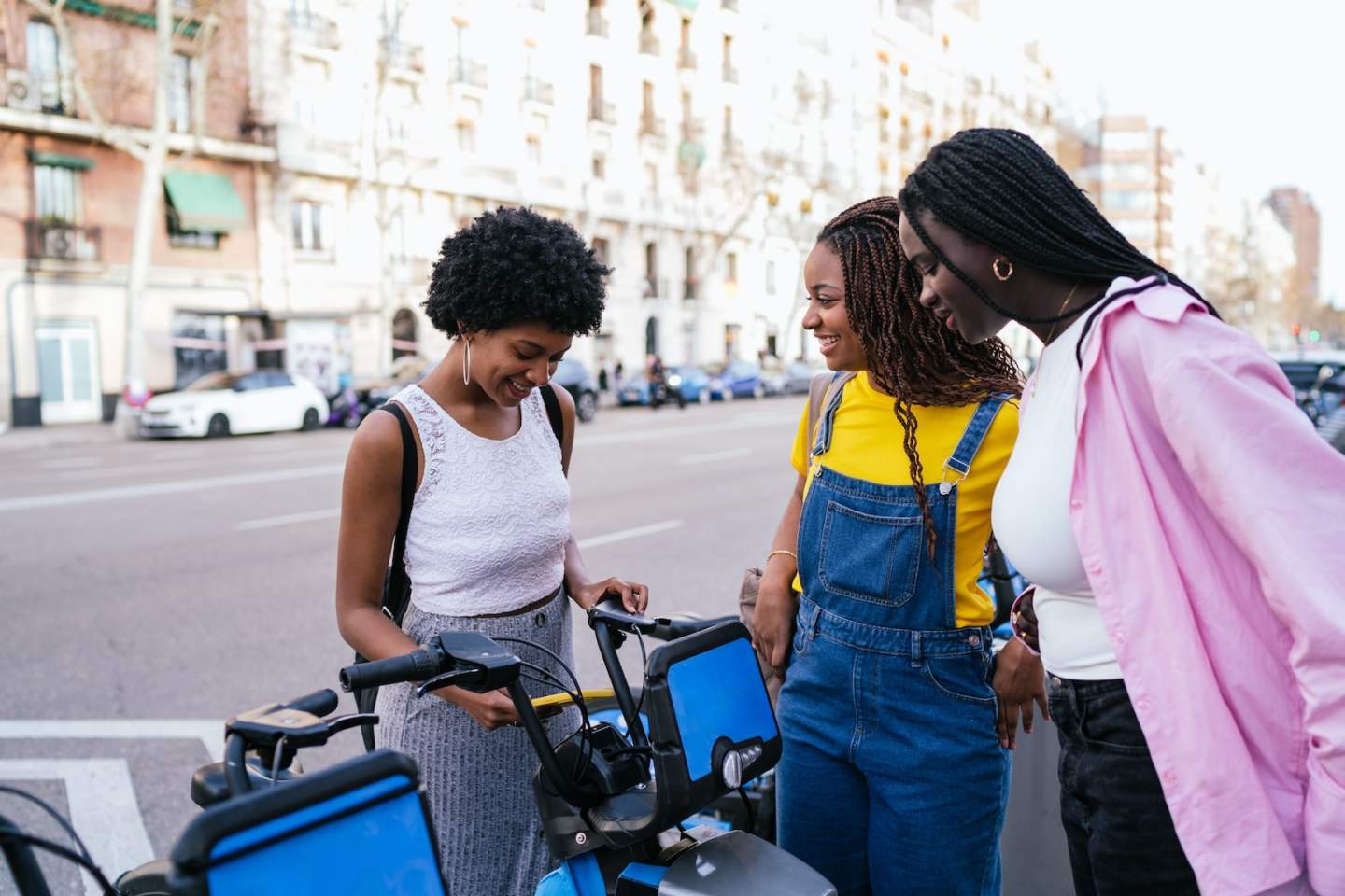 Three African women are using a smartphone to rent an electric bike on a city street, smiling and enjoying the moment.