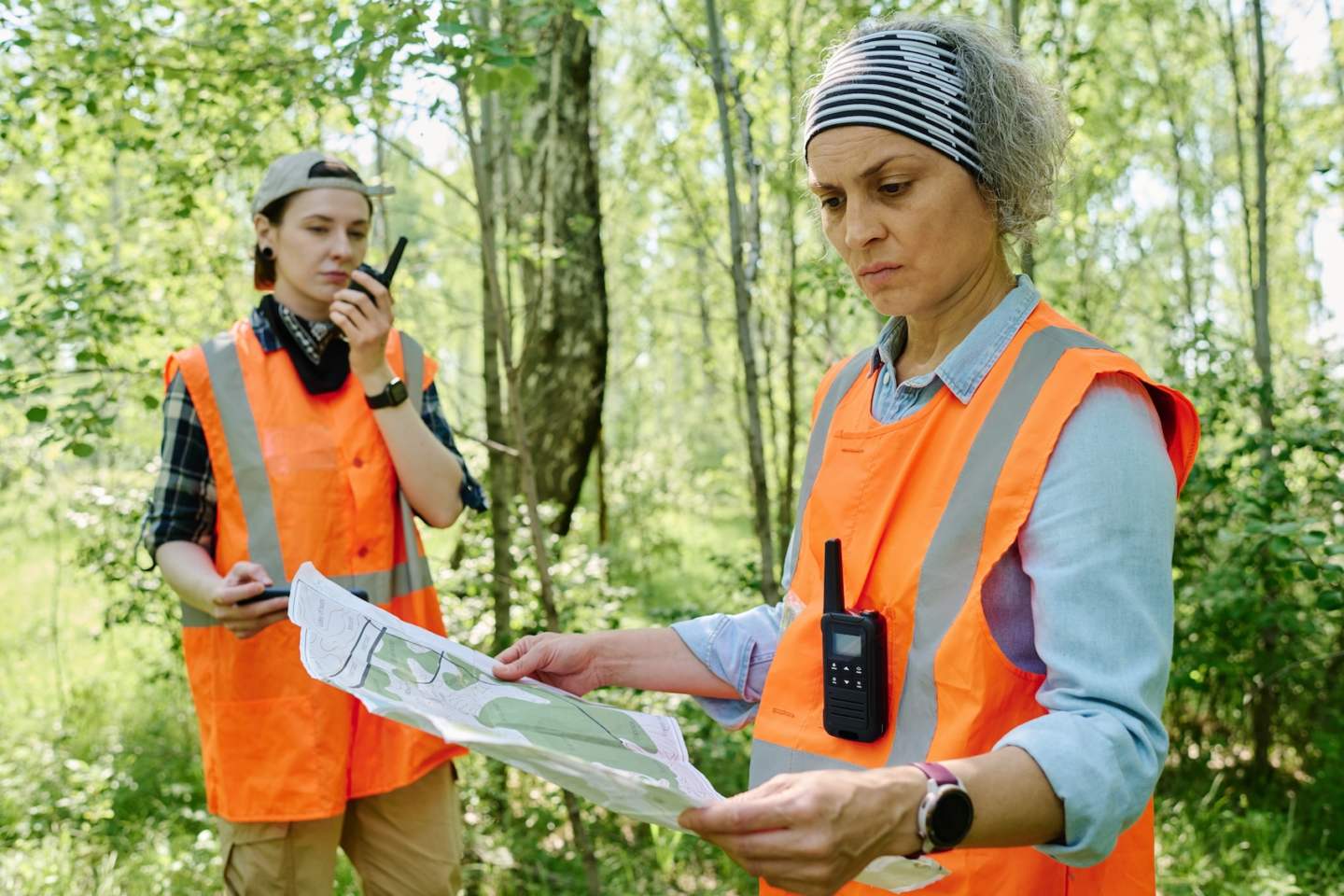 Mature serious female rescuer in headband and safety vest of acid orange color looking at unfolded map against young volunteer