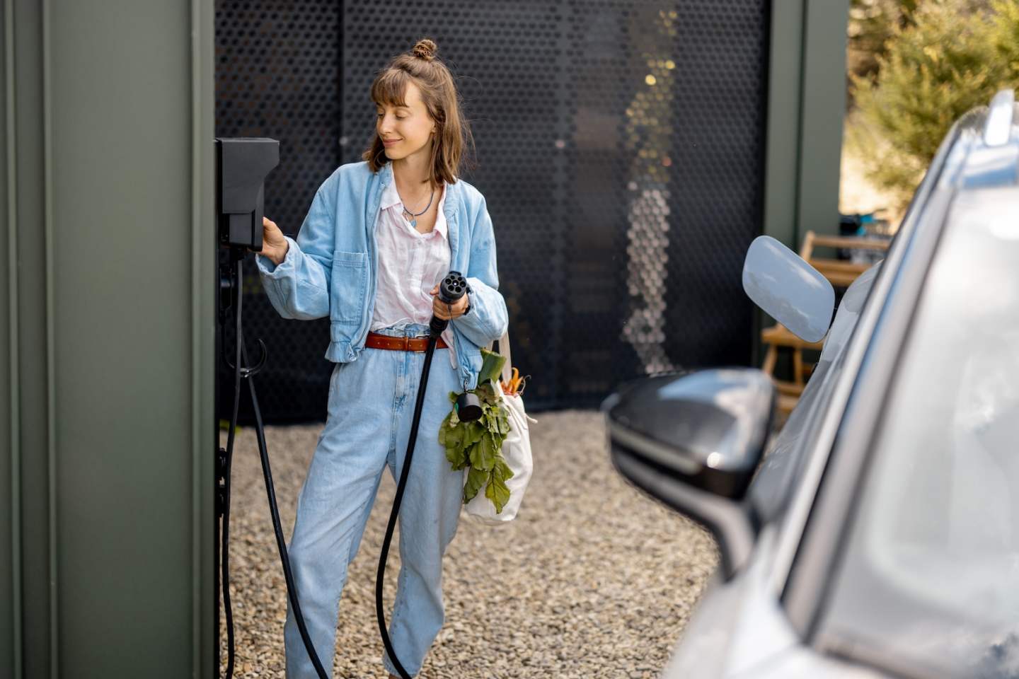 Young woman switches a car charger standing with bag full of fresh vegetables near her house. Concept of sustainable lifestyle and healthy food