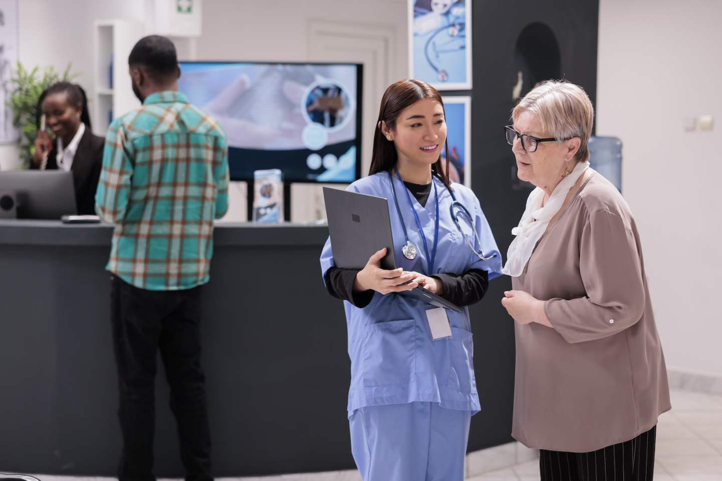 Asian geriatric specialist presenting results of routine tests to elderly caucasian patient. Senior woman looking medical images on computer at doctor office. Hospital admission area with diverse people.