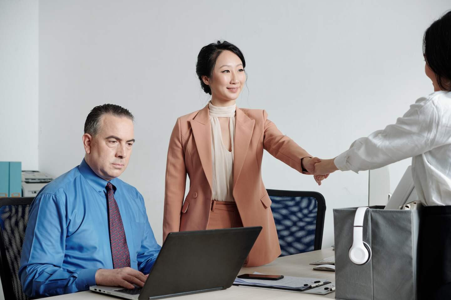 Serious mature entrepreneur working on laptop, his colleague greeting and shaking hand of new office worker