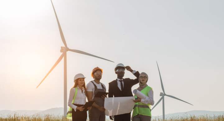 Multi ethnic group of engineers, technicians and managers in safety helmets having meeting outdoors about green energy. Men and women using tablet and drawing while working on windmill farm.