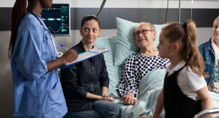 African american nurse monitoring sick elderly patient writing sickness expertise on clipboard during clinical recovery. Cheerful caring family visiting grandfather supporting him after surgery