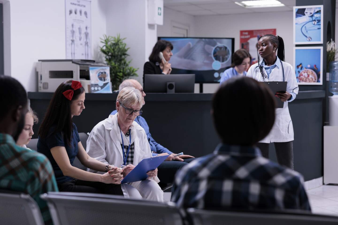 Senior pediatrician with clipboard doctor completing form for mother and child for routine checkup appointment in hospital reception. Diverse people waiting in modern private practice clinic.