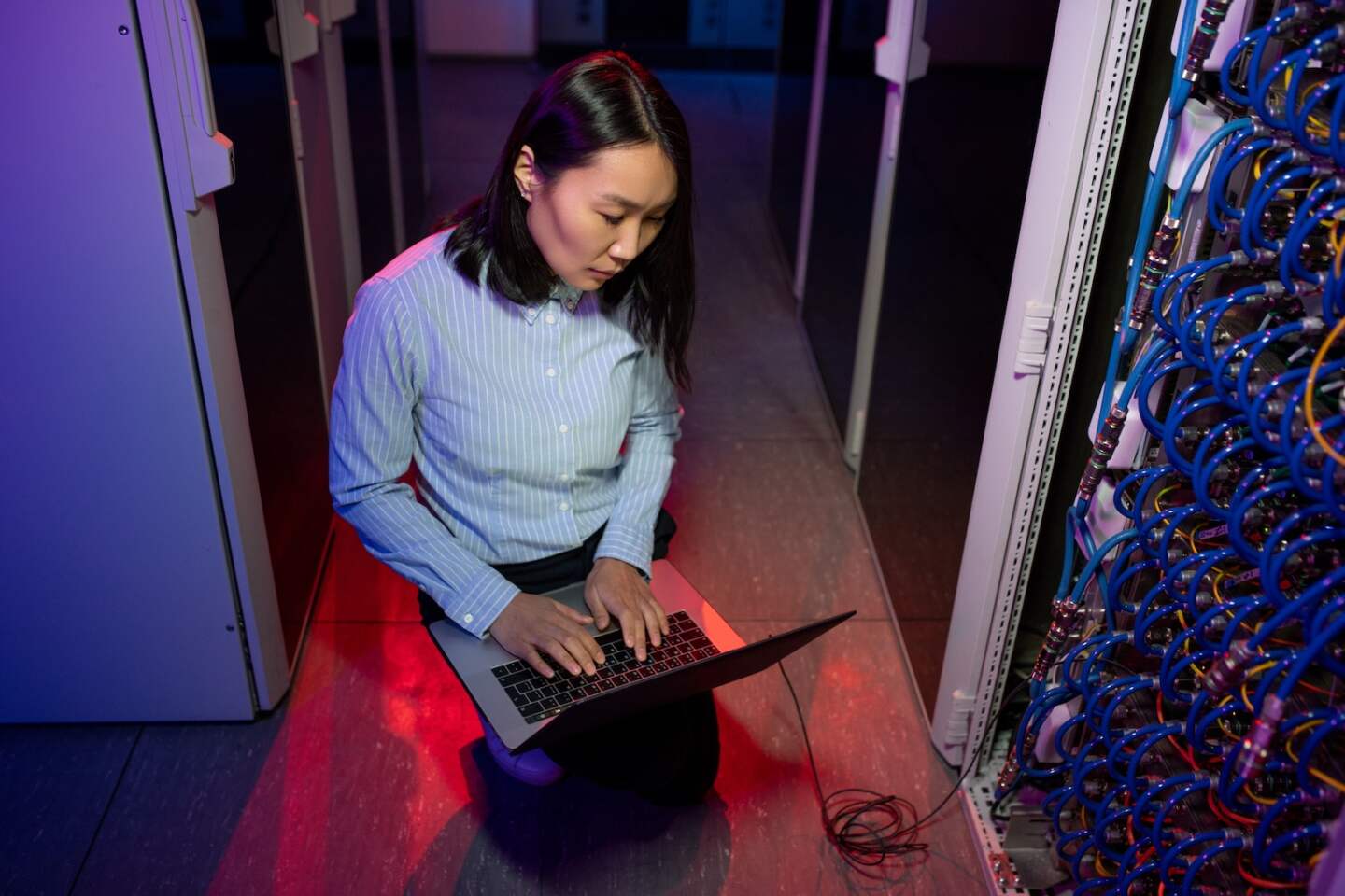 Above view of concentrated Asian IT engineer typing on laptop while connecting it to server in dark neon room