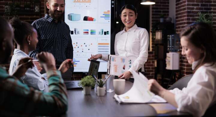 Startup business people talking and smiling at the end of late night sales meeting in corporate office. Marketing team casually discussing marketing strategy in front of wall screen tv with charts.